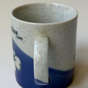May include: A ceramic mug with a speckled white and blue design. The mug features a blue wave pattern with a white flower graphic. The handle is white. The word "Carolina" is visible on the side of the mug.