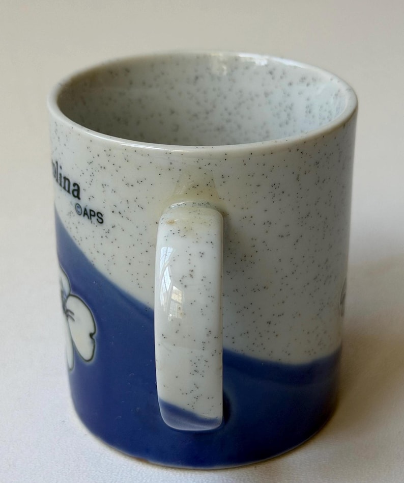 May include: A ceramic mug with a speckled white and blue design. The mug features a blue wave pattern with a white flower graphic. The handle is white. The word "Carolina" is visible on the side of the mug.