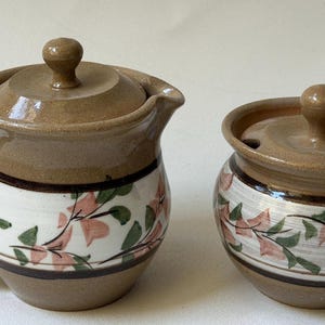 May include: A ceramic creamer with a handle and a matching sugar bowl with lids. Both pieces are tan with a white band featuring pink floral and green leaf designs, and brown trim.