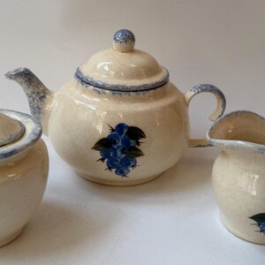 May include: A ceramic tea set consisting of a teapot with a lid, a sugar bowl with a lid, and a creamer pitcher. Each piece features a cream-colored base with a speckled texture, blue painted rims, and a cluster of blue berries with green leaves.