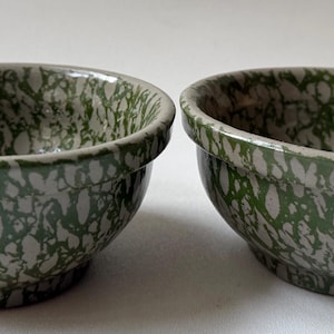 May include: Two small ceramic bowls featuring a speckled, mottled pattern in shades of forest green and off-white. Each bowl has a rounded shape with a distinct, slightly thickened rim and a smooth, glossy finish on the interior and exterior surfaces.