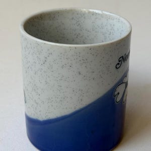 May include: A ceramic mug with a speckled white and blue design. The bottom half is solid blue, and the top half is white with black speckles. The mug has a floral design and the word "Nor" on it.