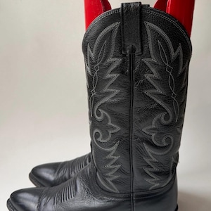Op de afbeelding: Een paar zwarte leren cowboylaarzen met decoratieve stiksels in een bloemmotief. De laarzen hebben spitse tenen en een gestapelde hak. Rode laarzenvormers zijn in de laarzen geplaatst.