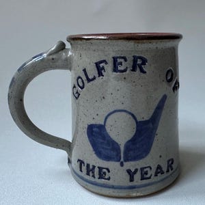 May include: A speckled ceramic mug with a brown rim and a curved handle. The mug features the words "GOLFER OF THE YEAR" in blue, along with a blue golf ball and tee design. The mug is a light gray color.