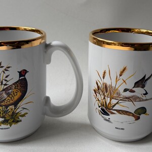 May include: Two white ceramic mugs with gold-toned rims. The left mug features a colorful illustration of a ring-necked pheasant with the text "RINGNECK PHEASANT" below. The right mug displays a mallard duck in water with the text "MALLARD" below.