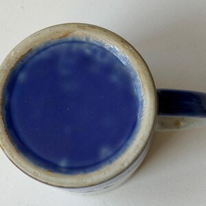 May include: A ceramic mug with a blue interior and handle. The exterior is a light beige with blue accents. The mug's base is a solid, glossy blue. The handle is a solid blue colour.