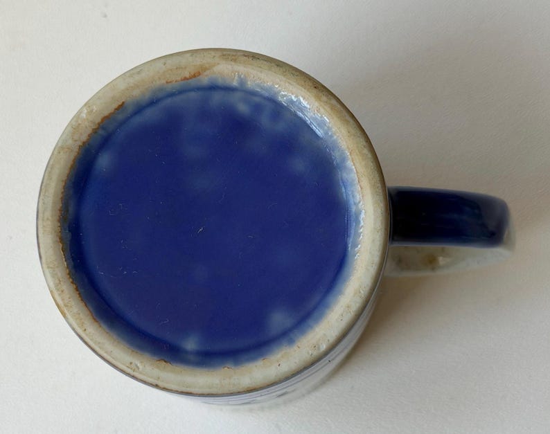 May include: A ceramic mug with a blue interior and handle. The exterior is a light beige with blue accents. The mug's base is a solid, glossy blue. The handle is a solid blue colour.