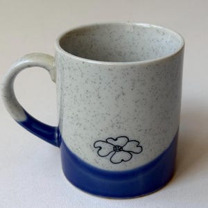 May include: A ceramic mug with a white speckled finish and a blue base. The mug features a black floral design and a curved handle. The handle and base are blue. The mug is approximately 10 cm tall.