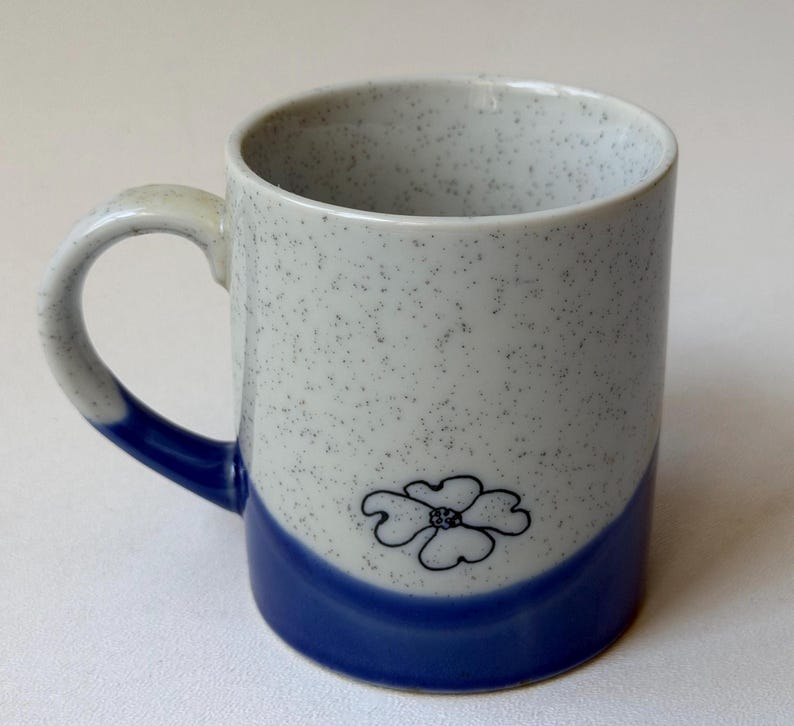 May include: A ceramic mug with a white speckled finish and a blue base. The mug features a black floral design and a curved handle. The handle and base are blue. The mug is approximately 10 cm tall.