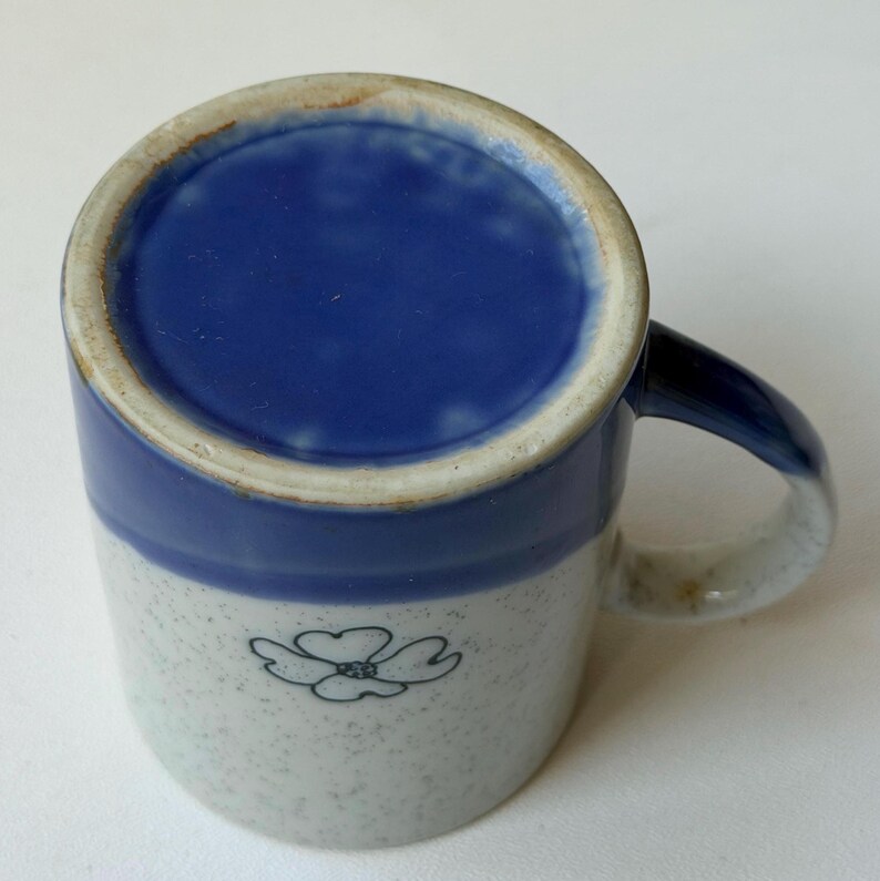 May include: A ceramic mug with a blue interior and a blue band around the top. The exterior is speckled white with a blue floral design. The handle is blue and white.