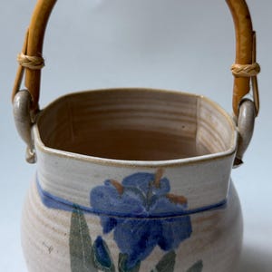 May include: A ceramic basket with a rounded body and a light beige glaze. The exterior features a hand-painted blue iris flower with green leaves and a horizontal blue line. It includes a curved, light brown bamboo handle attached to two ceramic loops.