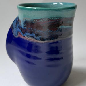 May include: A ceramic mug with a glossy finish, featuring a deep royal blue base that transitions into a dark purple and black band, topped with a bright turquoise rim. The mug has a distinct, rounded protrusion on one side instead of a handle.