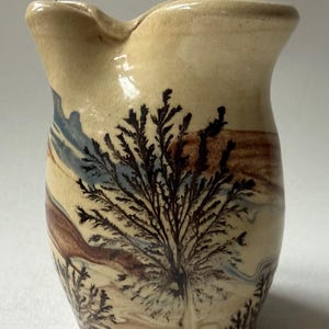 May include: A cream-colored ceramic pitcher featuring a prominent dark brown dendritic pattern resembling a tree or fern, accented by subtle horizontal bands of muted blue and earthy brown tones across the rounded body and a wide pouring spout.