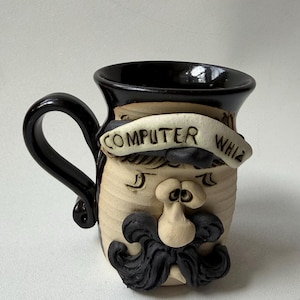 May include: A ceramic mug with a face design, featuring a black handle and rim. The mug has a tan base with a sculpted face, a large black mustache, and the text "COMPUTER WHIZ" on a banner.