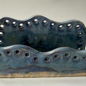 May include: A handcrafted ceramic desk organizer in shades of blue. The piece features a wavy design with multiple circular openings along the top edges. The base has a light beige color.