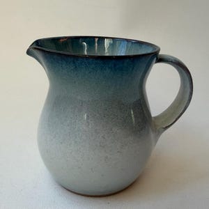 May include: A ceramic pitcher with a gradient color scheme, transitioning from white at the base to a deep blue at the rim. The pitcher has a rounded body, a curved handle, and a spout for pouring. The overall design is simple and elegant.