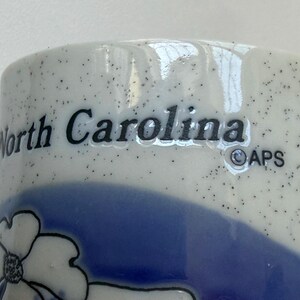 May include: Close-up of a ceramic mug with the words "North Carolina" in black lettering. The mug has a speckled white background with a blue floral design and a blue band. The APS logo is also visible.