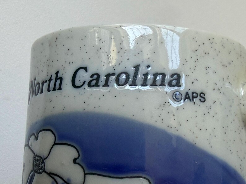 May include: Close-up of a ceramic mug with the words "North Carolina" in black lettering. The mug has a speckled white background with a blue floral design and a blue band. The APS logo is also visible.