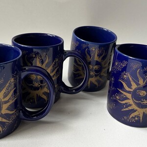 May include: Four navy blue ceramic mugs with a gold sun design. Each mug features a large sun with a face and radiating lines. The mugs have a curved handle and a glossy finish, ideal for tea or coffee.