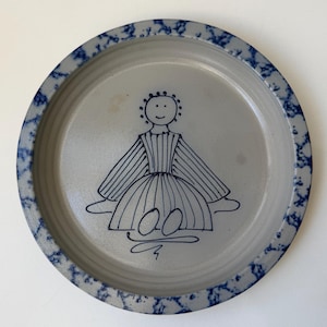Puede incluir: Un plato redondo de cerámica gris con un borde azul moteado. El plato presenta una ilustración dibujada a mano de una figura con un vestido. La figura está dibujada en azul, con una cara sonriente y un vestido a rayas.