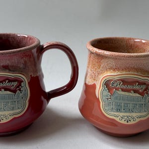 May include: Two ceramic mugs with a rounded shape and a handle on the right side. Each mug features a cream-colored rim that transitions into a solid red or orange base. A central label displays the text Bloomsbury above a blue house illustration.