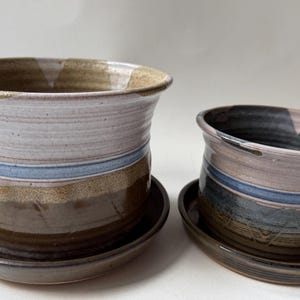 May include: Two ceramic planters with matching saucers, featuring a layered glaze design in shades of cream, light blue, and dark brown. The larger planter on the left and the smaller one on the right both have a wide, flared rim and a cylindrical body.