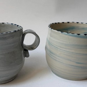 May include: Two handmade ceramic mugs. The mugs are gray and off-white with a swirl pattern. Each mug has a rounded body, a handle, and a decorative rim with small black accents. The mugs are suitable for coffee, tea, or other beverages.