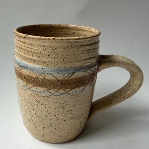 May include: A speckled beige ceramic mug featuring a cylindrical body with a wide handle. The exterior displays a horizontal band with a light blue stripe and a brown, hand-drawn, intertwined line pattern. The rim has subtle horizontal ridges.