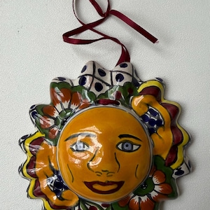 May include: A ceramic sun-shaped ornament with a smiling face, painted in shades of yellow, orange, blue, green, and red. The sun has a decorative border and a maroon ribbon for hanging. A cheerful, handcrafted decoration.