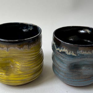 May include: Two handmade ceramic cups with a unique, textured design. One cup is yellow, the other is blue, both with dark rims and interiors. The cups have a wavy, ridged exterior, adding visual interest to the functional pottery.