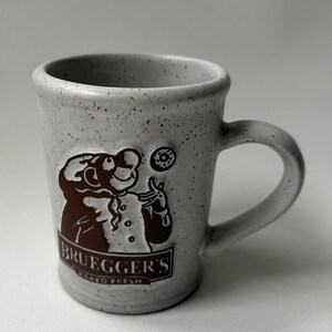 May include: A speckled gray ceramic mug with a brown graphic of a chef juggling a donut. The mug has a curved handle and the text "BRUEGGER'S BAKED FRESH" is printed on the front.
