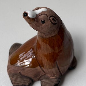 May include: A brown ceramic seal figurine with a glossy finish. The seal has a white marking on its head and black eyes. The figurine is sitting on its belly with its flippers visible. The overall design is simple and charming.