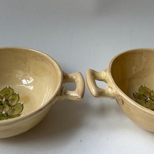 May include: Two cream-colored ceramic bowls with handles. Each bowl features a green leaf design in the center. The bowls have a rounded shape and are suitable for serving soups or stews.