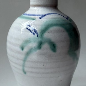 May include: A small, off-white ceramic vase with a flared rim and a bulbous body. The surface features horizontal ridges and hand-painted brushwork in muted green and dark blue, creating a simple, organic pattern across the glazed exterior.