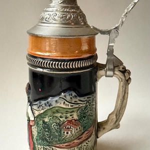 May include: A ceramic beer stein featuring a metallic lid with a pointed finial and a hinged thumb lift. The body is dark blue with a raised, colorful relief depicting a landscape with a house, trees, and a person, framed by orange bands.