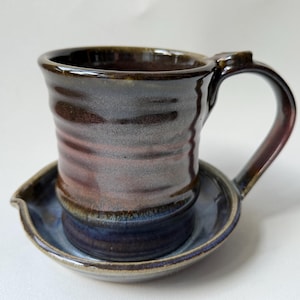 Puede incluir: Taza de cerámica hecha a mano con platillo a juego. La taza presenta un esmalte marrón oscuro, gris y marrón rojizo con un asa curva. El platillo es azul con un borde ligeramente elevado. Un juego de vajilla artesanal único.