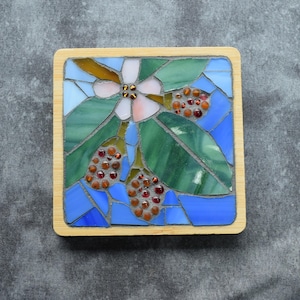May include: A square coaster featuring a mosaic design of a white flower with green leaves and brown berries. The mosaic is composed of various shades of blue, green, and white glass pieces set in a light-colored wooden frame with rounded corners.