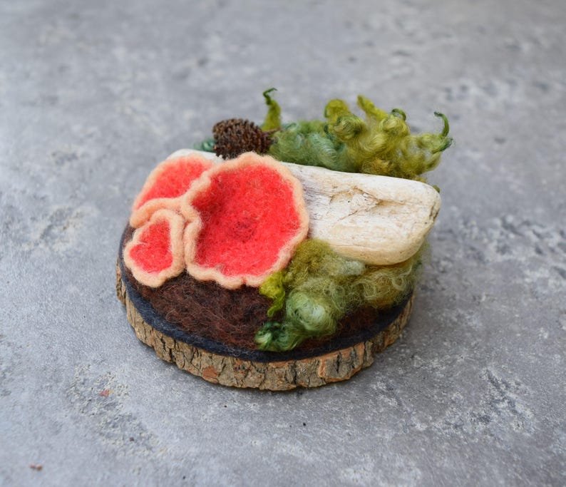 May include: A handcrafted felted mushroom sculpture with vibrant red caps and beige edges, set on a wooden slice base. The artwork includes green moss and a piece of driftwood, creating a miniature forest scene.