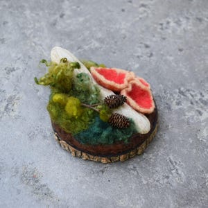 May include: A handcrafted felted brooch featuring a miniature forest scene. The design includes red mushroom caps, green moss, a white log, and small pinecones, all set on a wooden base. The brooch is a unique, nature-inspired accessory.