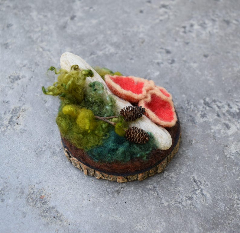 May include: A handcrafted felted brooch featuring a miniature forest scene. The design includes red mushroom caps, green moss, a white log, and small pinecones, all set on a wooden base. The brooch is a unique, nature-inspired accessory.