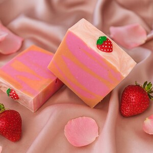 May include: Two square soaps with pink and orange swirls, topped with a strawberry embellishment. Fresh strawberries and pink rose petals are scattered around the soaps on a pink satin surface. The soaps are a handmade, decorative item.