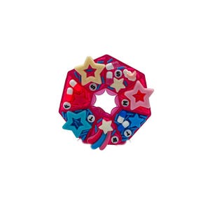 May include: A bright pink, star-shaped charm with a central hole. The charm is decorated with various colourful stars, a shooting star, and small, white, cylindrical shapes. The design is set against a white background.