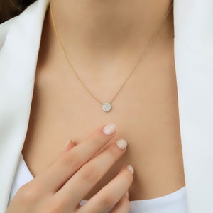 May include: A delicate gold necklace with a small, round pendant set with clear gemstones. The pendant hangs from a fine gold chain, creating a minimalist and elegant design. The necklace is shown against a white background.