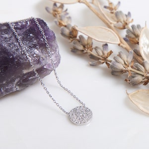May include: A silver necklace featuring a round pendant encrusted with small diamonds. The necklace is displayed on a white surface, next to a purple amethyst and dried lavender. The pendant is about 1.3 cm in diameter.