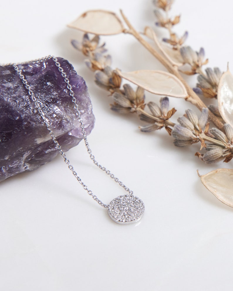 May include: A silver necklace featuring a round pendant encrusted with small diamonds. The necklace is displayed on a white surface, next to a purple amethyst and dried lavender. The pendant is about 1.3 cm in diameter.