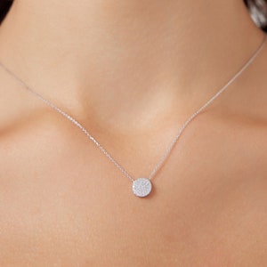 May include: A delicate silver necklace with a small, round pendant set with sparkling, clear gemstones. The pendant hangs from a thin chain, creating a minimalist design. The necklace is displayed against a skin tone background.