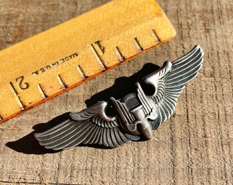 Puede incluir: Un pin de alas militares de color plateado con un diseño de avión estilizado. El pin está sobre una superficie de madera con una regla que dice "Made in U.S.A."