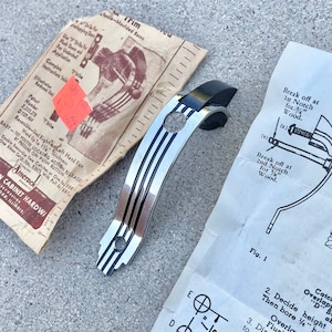 May include: A silver and black cabinet door handle with a black plastic backplate. The handle has a grooved design and a small hole for a screw. The instructions for installing the handle are visible in the background.