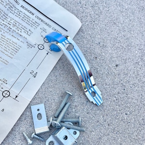 May include: A silver cupboard catch with a blue plastic plunger and instructions for installation. The instructions include a diagram showing the placement of the catch and screws. The image also shows the hardware needed for installation, including screws, a metal plate, and a small metal pin.
