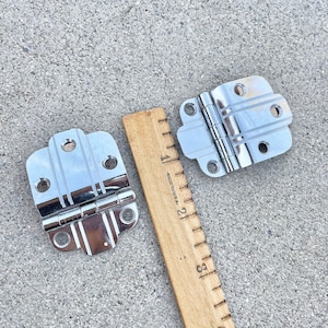 May include: Two silver-toned door hinges with a decorative design. The hinges are 5 cm long and are shown next to a wooden ruler.